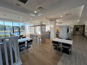 Un vaste espace intérieur doté d’un plancher en bois, de tables et de chaises blanches, et de grandes fenêtres donnant sur une piscine extérieure.