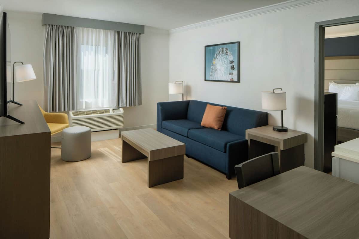 Una habitación de hotel moderna con sofá, dos mesas auxiliares, una mesa de centro, una silla, un escritorio y una cama.