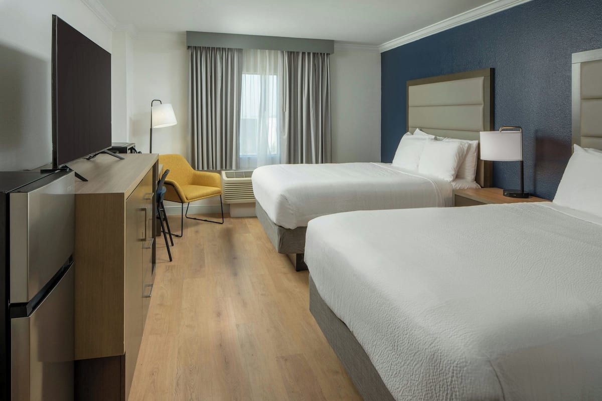 Habitación de hotel con dos camas, un escritorio con televisor, una silla y una ventana con cortinas.
