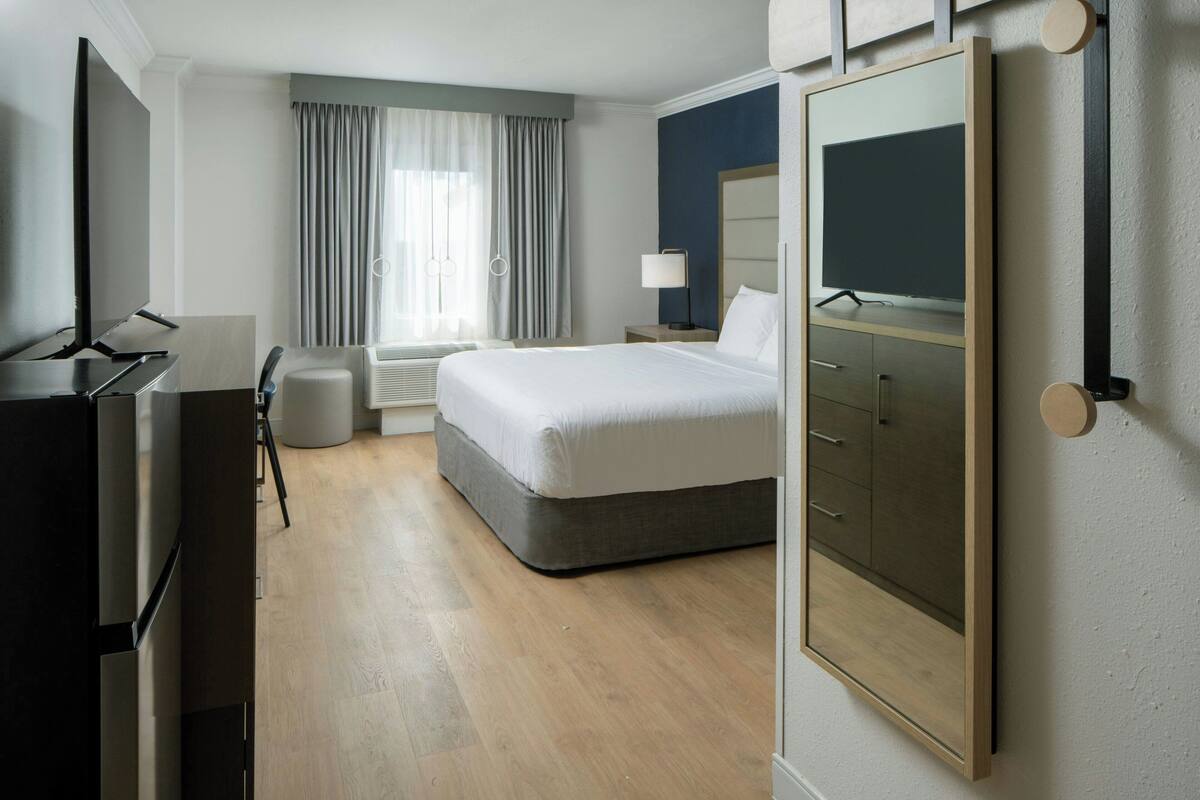 Una habitación de hotel con cama, escritorio con computadora, televisor, armario y ventana con cortinas.