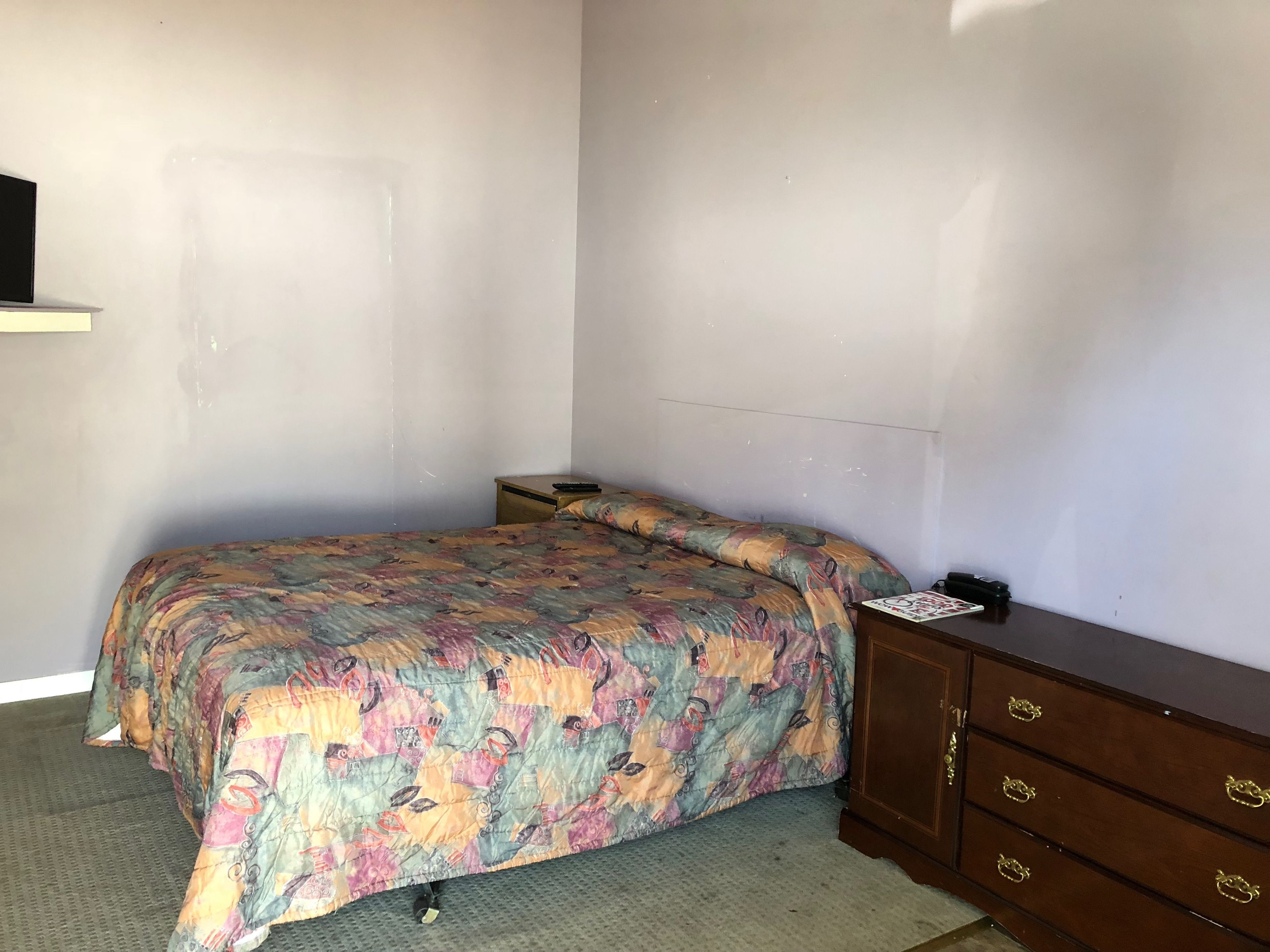 A small, sparsely furnished room with a bed, a nightstand, a dresser, and a door.