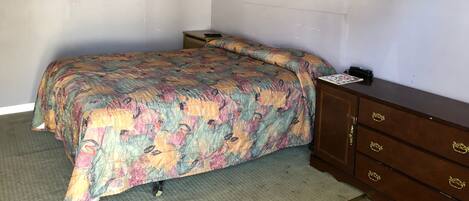 A small, sparsely furnished room with a bed, a nightstand, a dresser, and a door.