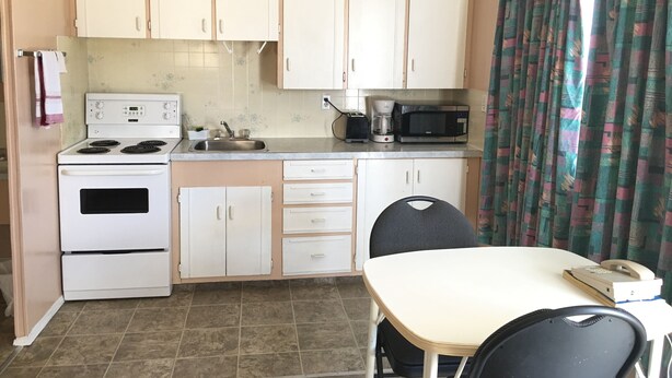 A compact kitchen with a stove, sink, microwave, and a small dining area with a table and chairs. - Bluebird Motel (Innisfail)