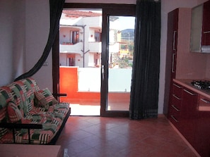 A living room with a sofa, a kitchen area with cabinets, and a sliding glass door leading to a balcony. - Residence Ibiscus (Castelsardo)