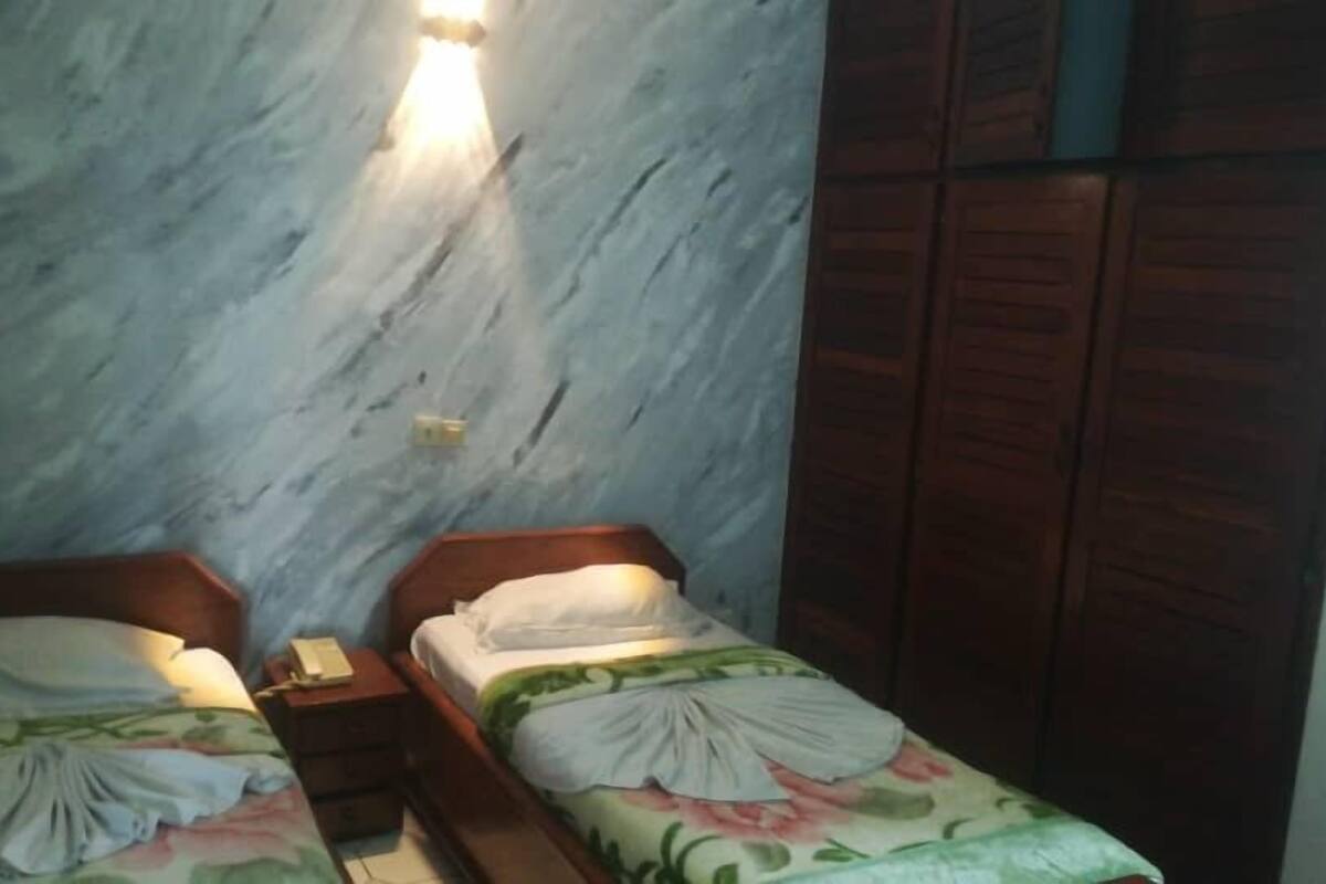 A bedroom with two single beds, wooden headboards, a nightstand with a lamp, and a wall-mounted light fixture.