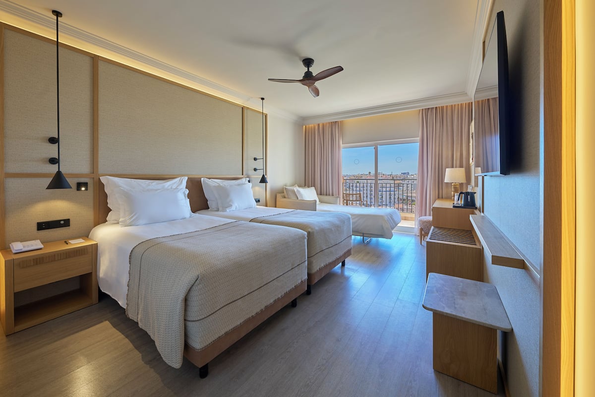 A hotel room with two beds, a desk, a TV, and a large window with a city view.