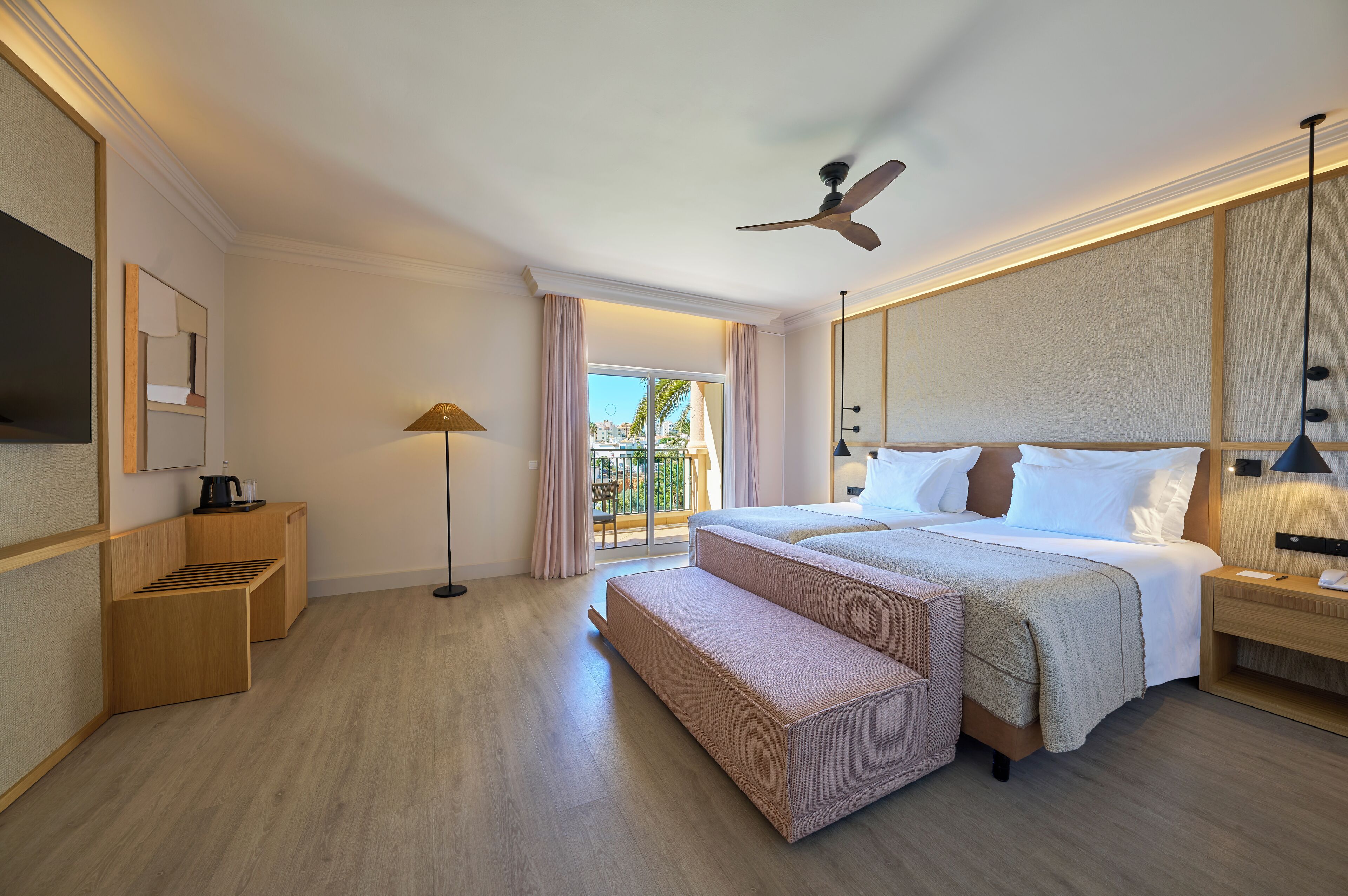 A hotel room with a bed, a bench, a ceiling fan, a TV, a lamp, and a view of the outdoors.