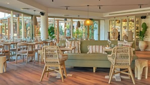 Un restaurante espacioso con suelo de madera, sillas de mimbre, un sofá verde y grandes ventanales con vistas al exterior.