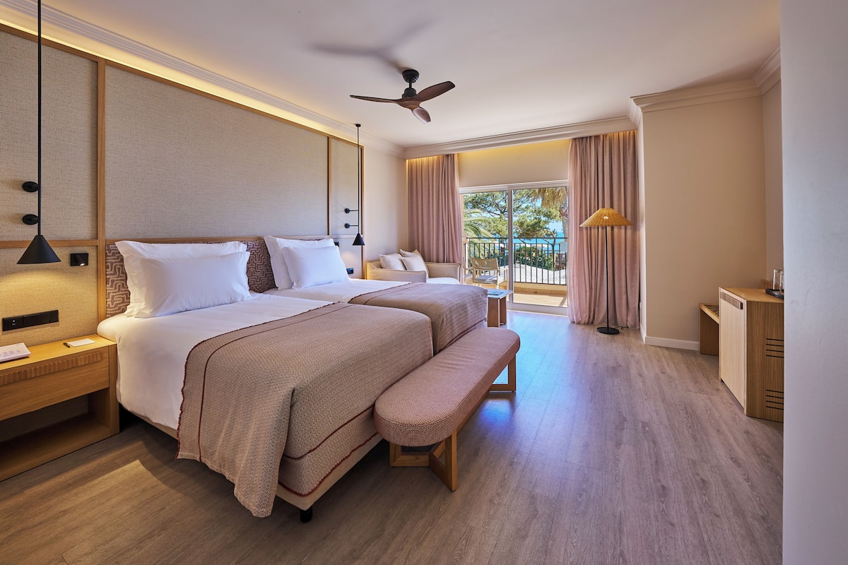A hotel room with two beds, a bench, a desk, a ceiling fan, and a view of the outdoors.