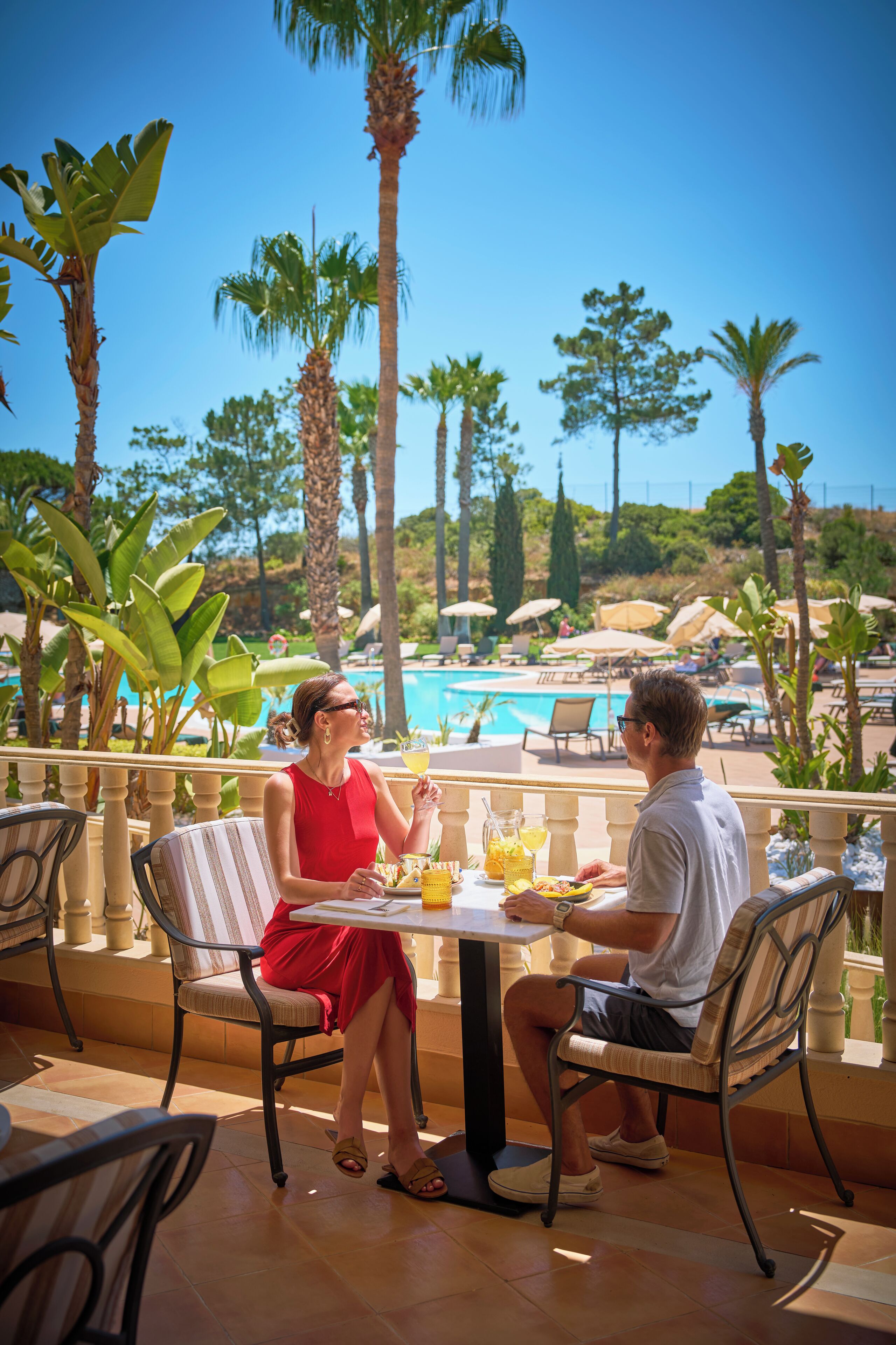 Una pareja cenando en una mesa al aire libre con vistas a una piscina, rodeada de palmeras y sillones de descanso.