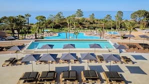 Zona de piscina en un complejo turístico, con tumbonas y sombrillas, rodeada de palmeras y vistas al mar.