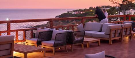 A deck with outdoor seating and a view of the ocean at sunset.