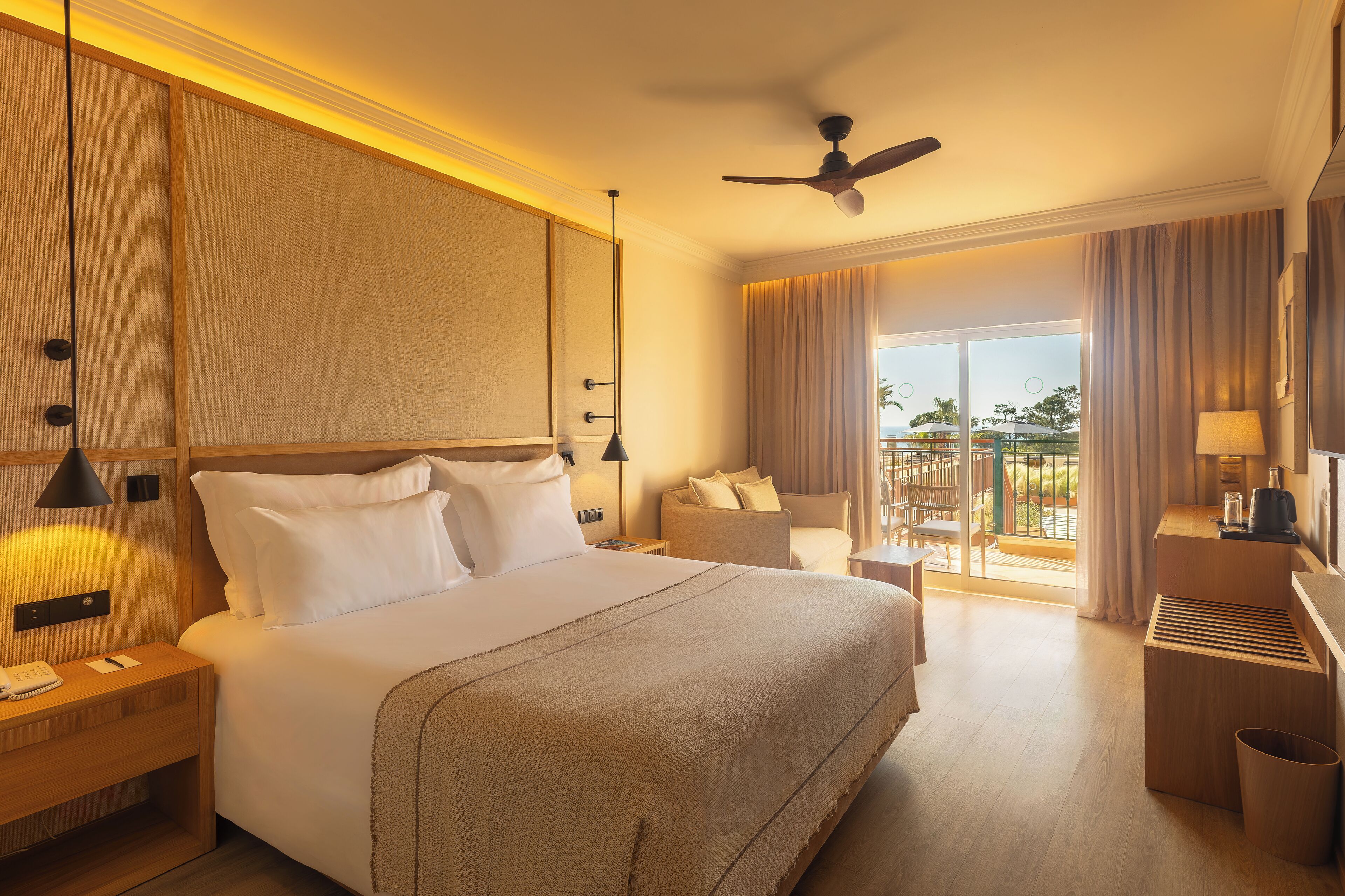 A hotel room with a large bed, a desk with a lamp, a ceiling fan, and a view of the outdoors.