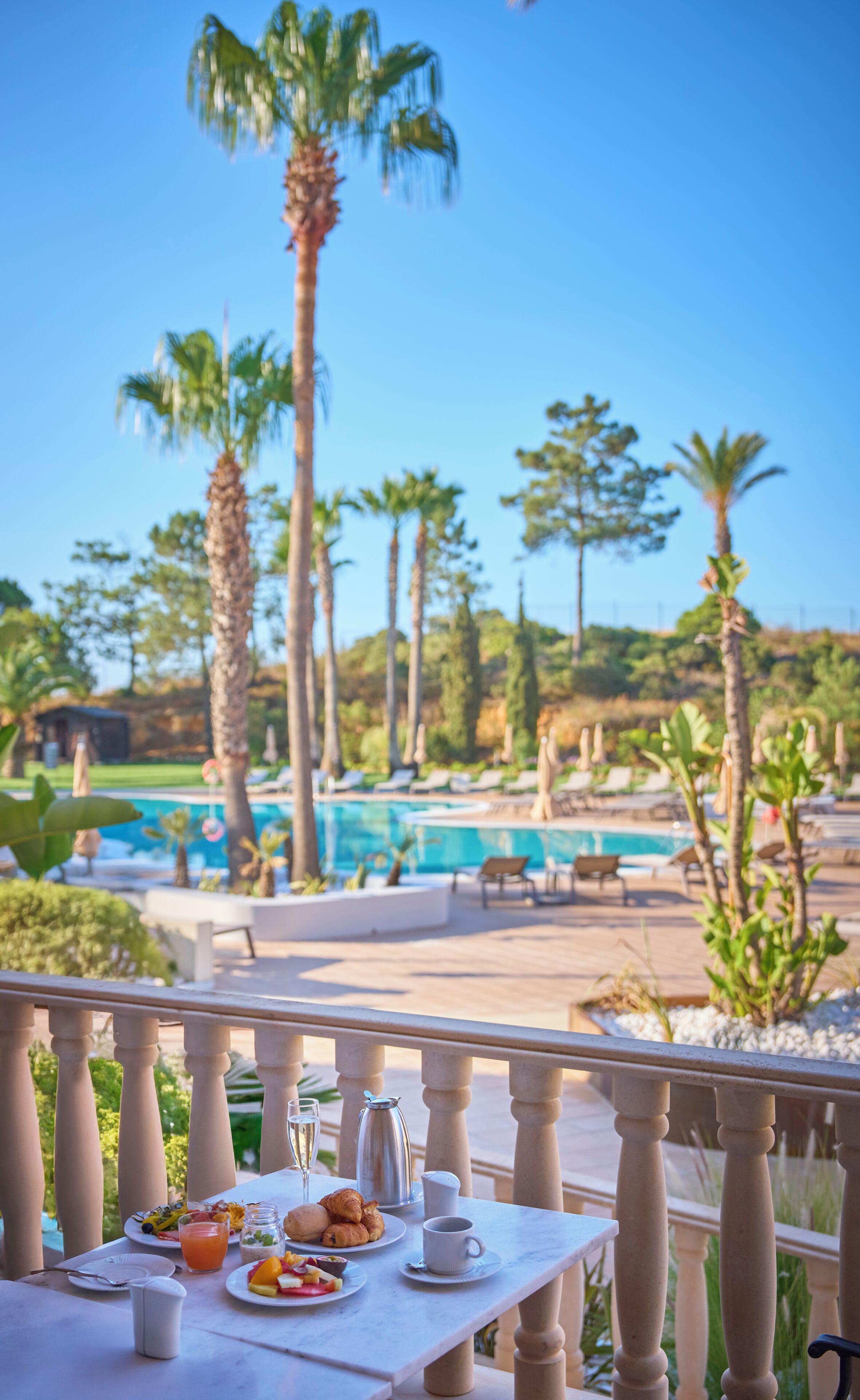 Un tavolo da pranzo con cibi per la colazione, con vista su una zona piscina con palme e poltrone relax.