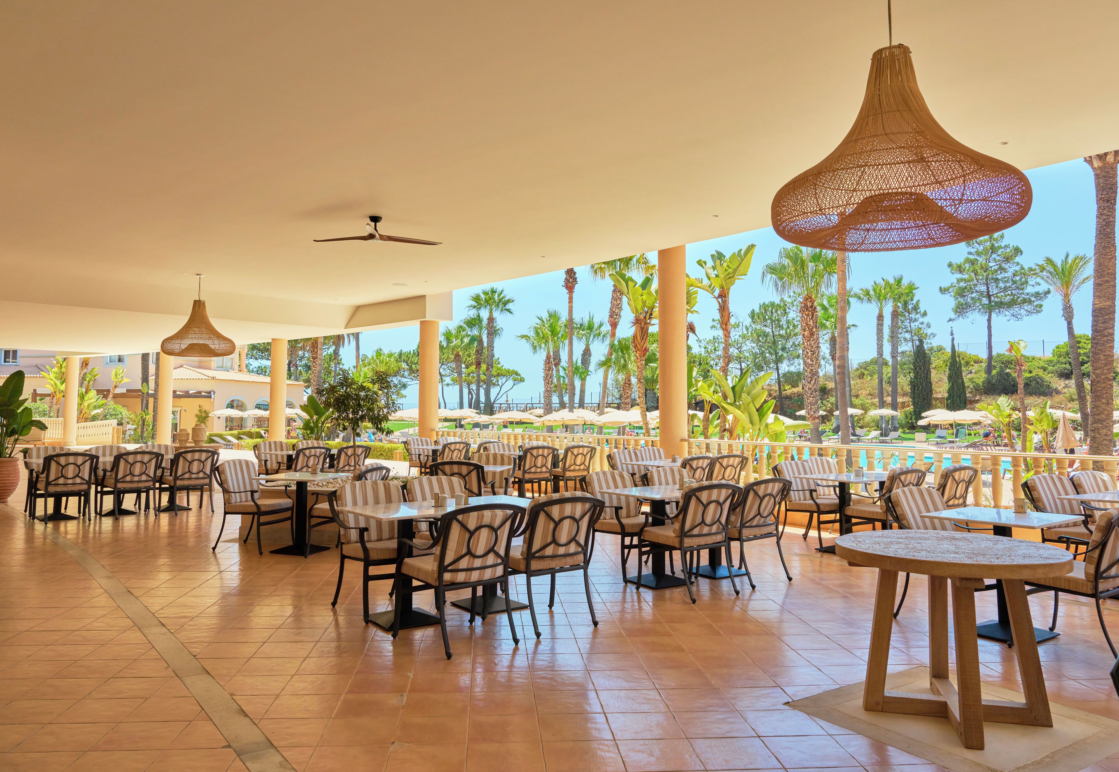 A spacious outdoor dining area with multiple tables and chairs, large hanging lights, and a view of palm trees and a pool.