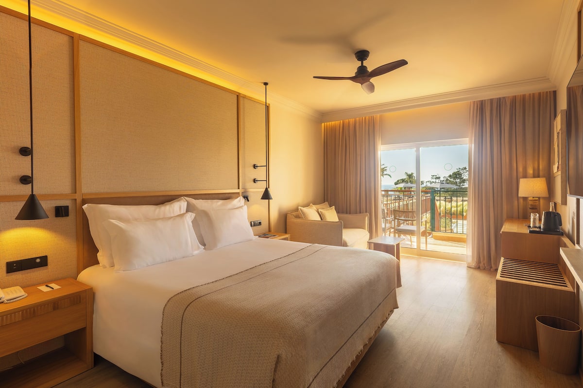 A hotel room with a large bed, a desk with a lamp, a ceiling fan, and a view of the outdoors.