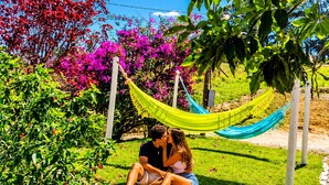 Una pareja sentada sobre la hierba bajo una hamaca, rodeada de exuberante follaje y plantas con flores.