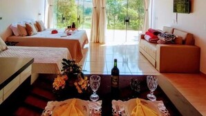 Una habitación con techo de madera, cama, sofá, mesa de comedor con una botella y vasos, y vista a través de amplios ventanales.