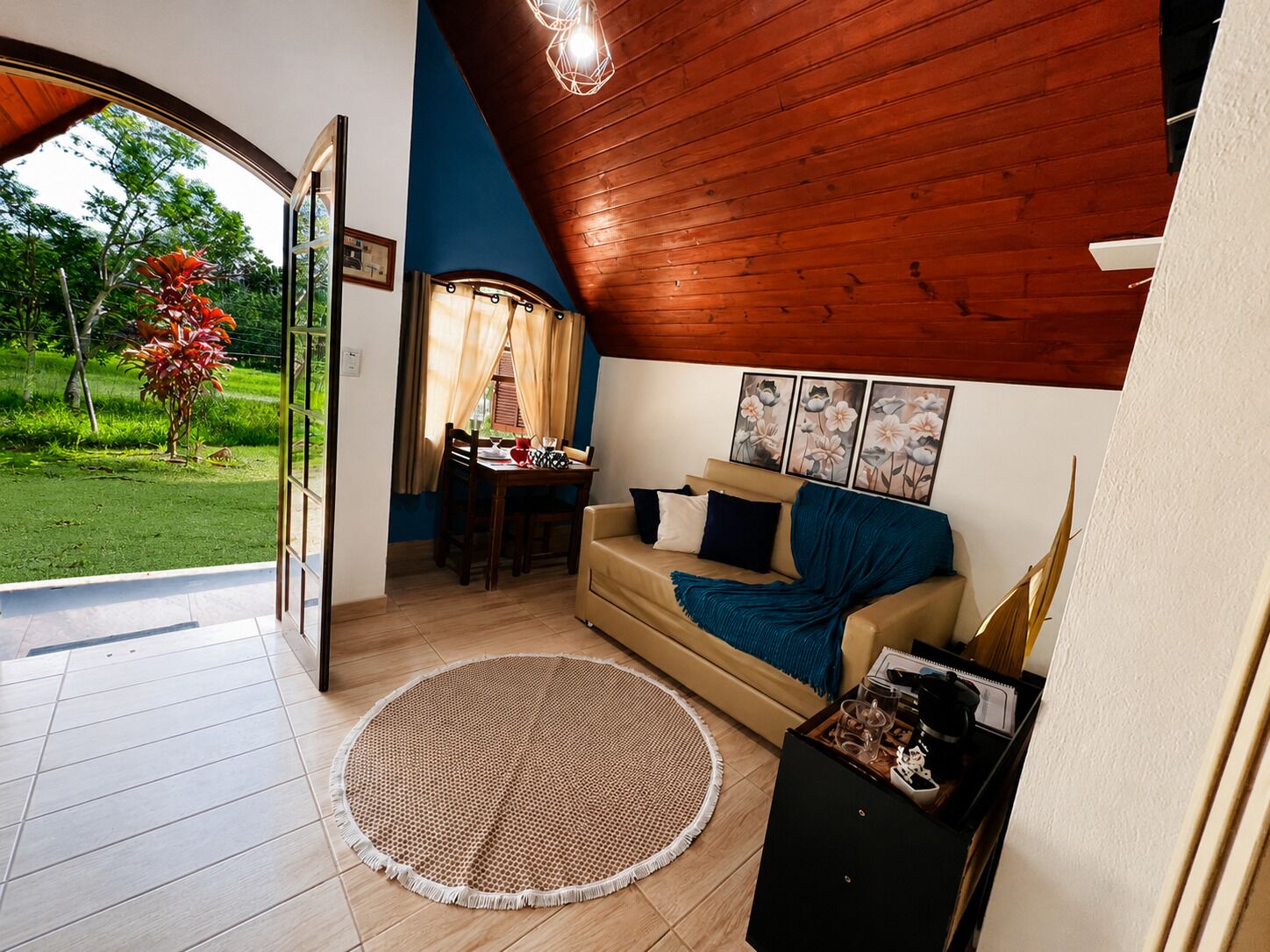 Een gezellige zithoek met een houten plafond, een rond geweven vloerkleed, een bank met kussens en een eettafel met stoelen.