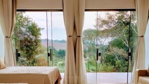 Una habitación con cama, una mesa de comedor con servilletas amarillas y vistas a la vegetación a través de grandes ventanales.