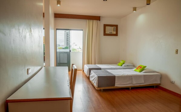 Een hotelkamer met houten vloer, twee enkele bedden met groene kussens, een kleine tafel en een raam met gordijnen.