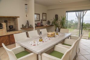 Un comedor con una mesa blanca, sillas de mimbre con cojines verdes, un camino de mesa decorativo y vistas al exterior a través de un ventanal.