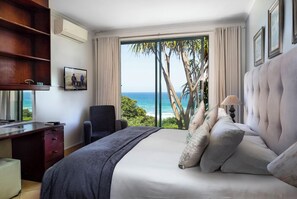 Un dormitorio con una cama grande, un escritorio y una silla. Hay una ventana con vista al océano y palmeras.