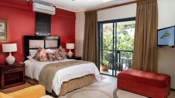 A hotel room with a red accent wall, a bed with floral pillows, a red ottoman, a TV, and a balcony with a view of greenery.