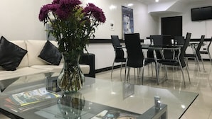 A modern hotel lobby with a glass coffee table, a white sofa with black cushions, a vase with purple flowers, and a dining area with black chairs and a table. - Hotel Ejecutivo Embajada (Bogotá)