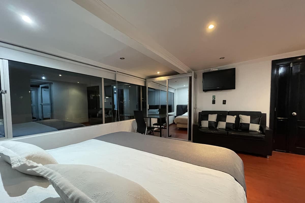 A modern hotel room with a large bed, a seating area with a table and chairs, a sofa, and a flat-screen TV mounted on the wall.
