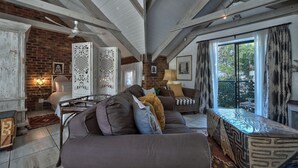 A cozy living room with a brown sofa, a patterned ottoman, and a brick wall. There is a ceiling fan, a window with curtains, and a small table with a lamp.