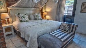 A bedroom with a large bed, a bedside table with a lamp, a bench with a cushion, and a view of the outside.
