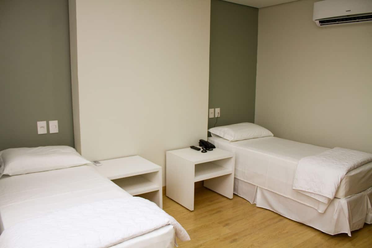 A small, clean room with two single beds, a bedside table with a phone, and an air conditioning unit on the wall.