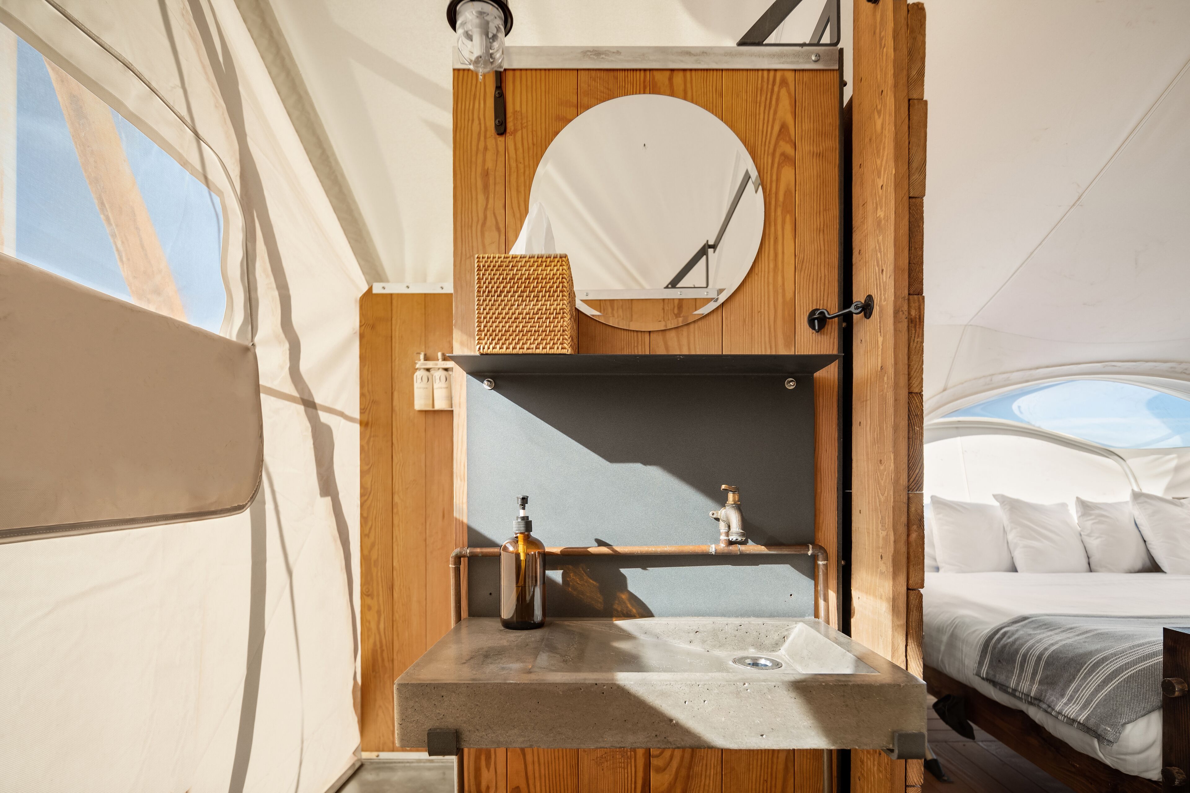A compact, well-lit interior with a wooden vanity, a round mirror, a small sink, and a bed with a view of the sky.