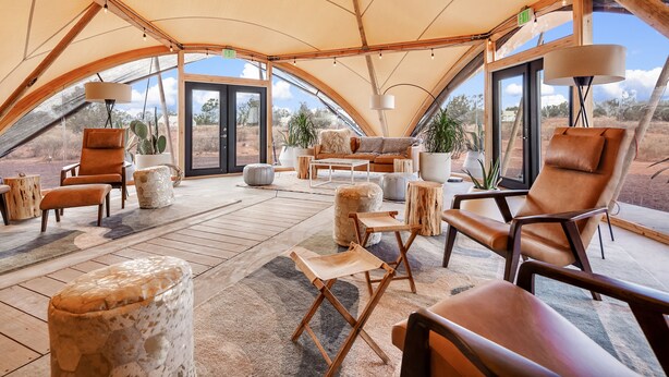 Un hébergement spacieux sous tente, doté d’un espace salon central, de meubles en bois et de grandes fenêtres offrant une vue sur le paysage environnant.