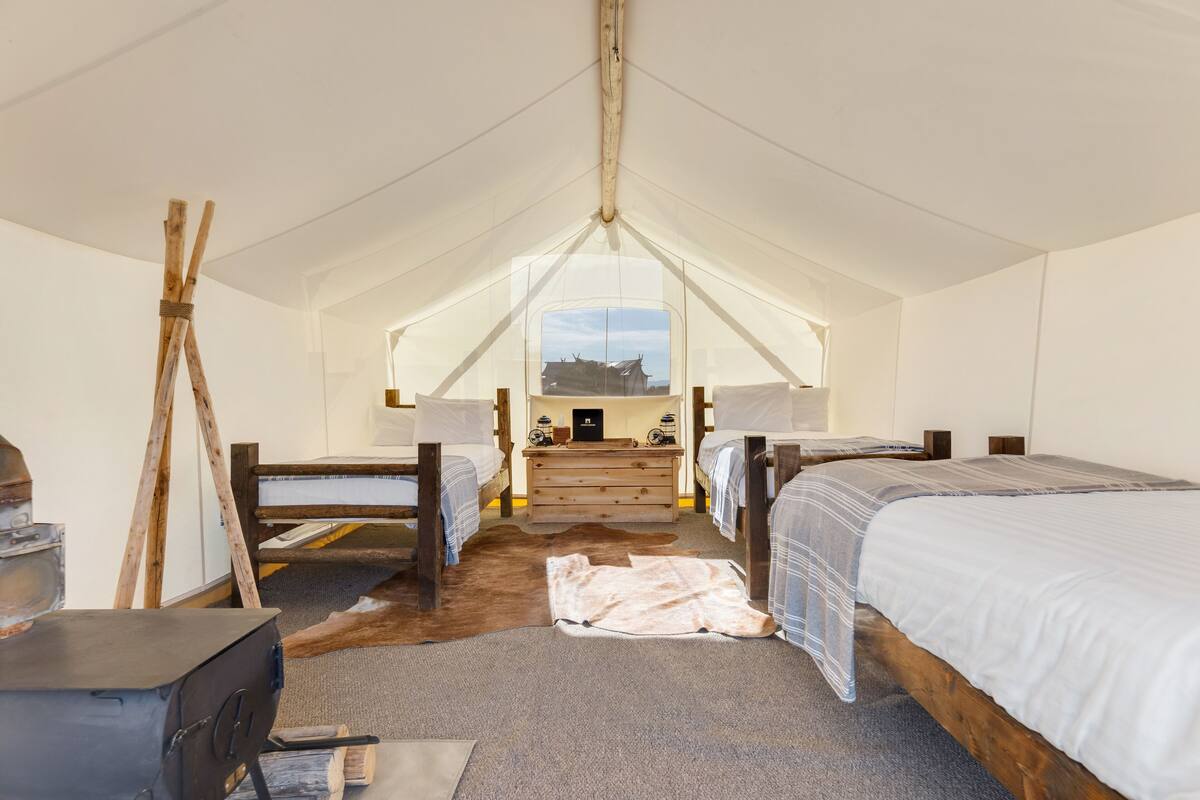 Une pièce aménagée avec une tente, comprenant deux lits, une commode en bois, un poêle et un tapis en peau de vache.