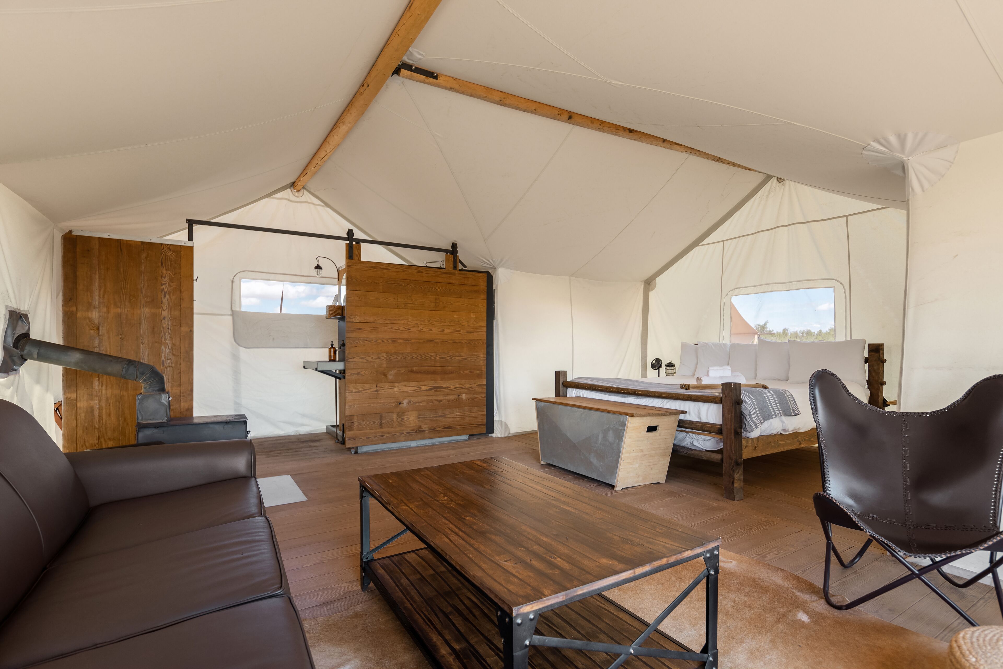 A tented accommodation with a wooden table, leather sofa, and a bed with a wooden frame.