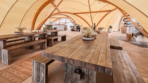 Une tente spacieuse, dotée de bancs et de tables en bois, de plantes en pot et d’un espace central avec une longue table.