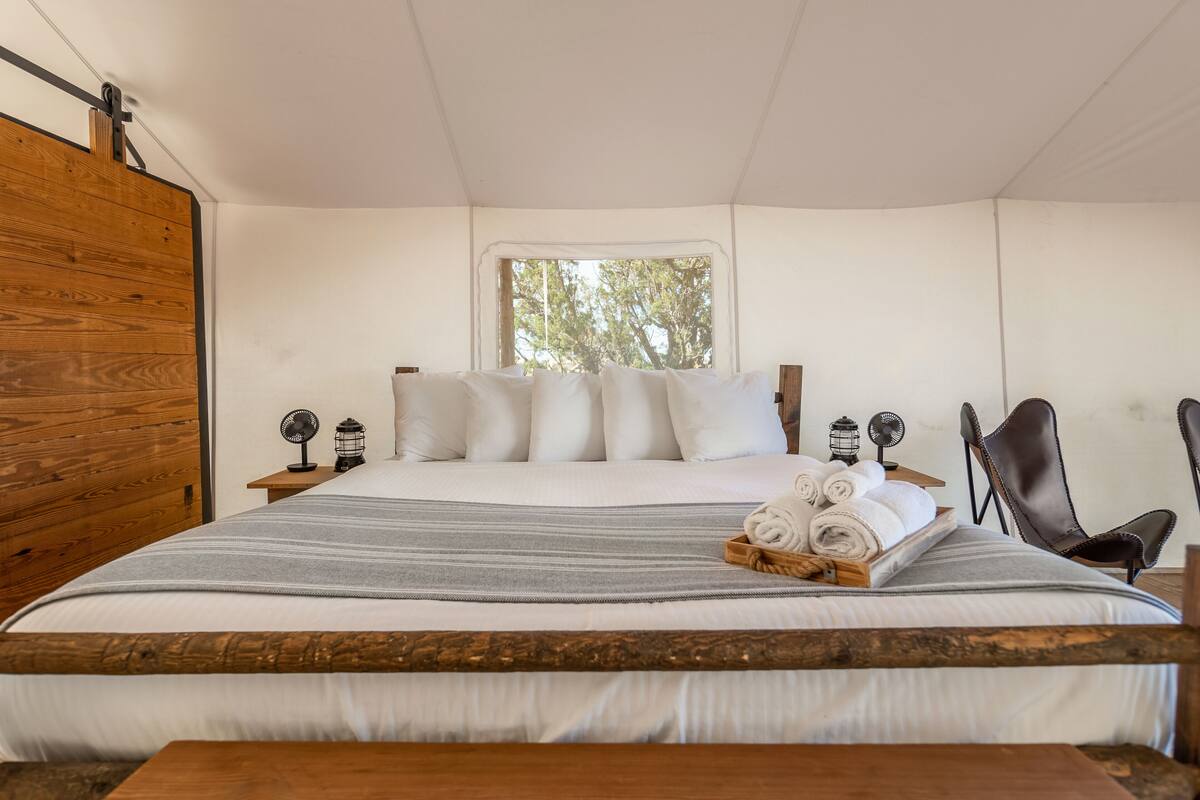 Un lit bien fait, avec du linge de lit blanc, une couverture à rayures grises et une tête de lit en bois. Il y a deux tables de chevet avec des ventilateurs et des lanternes, ainsi qu’une chaise avec un siège en cuir.