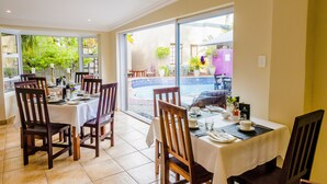 A dining area with tables set for four, large windows, and a view of a pool and greenery outside.