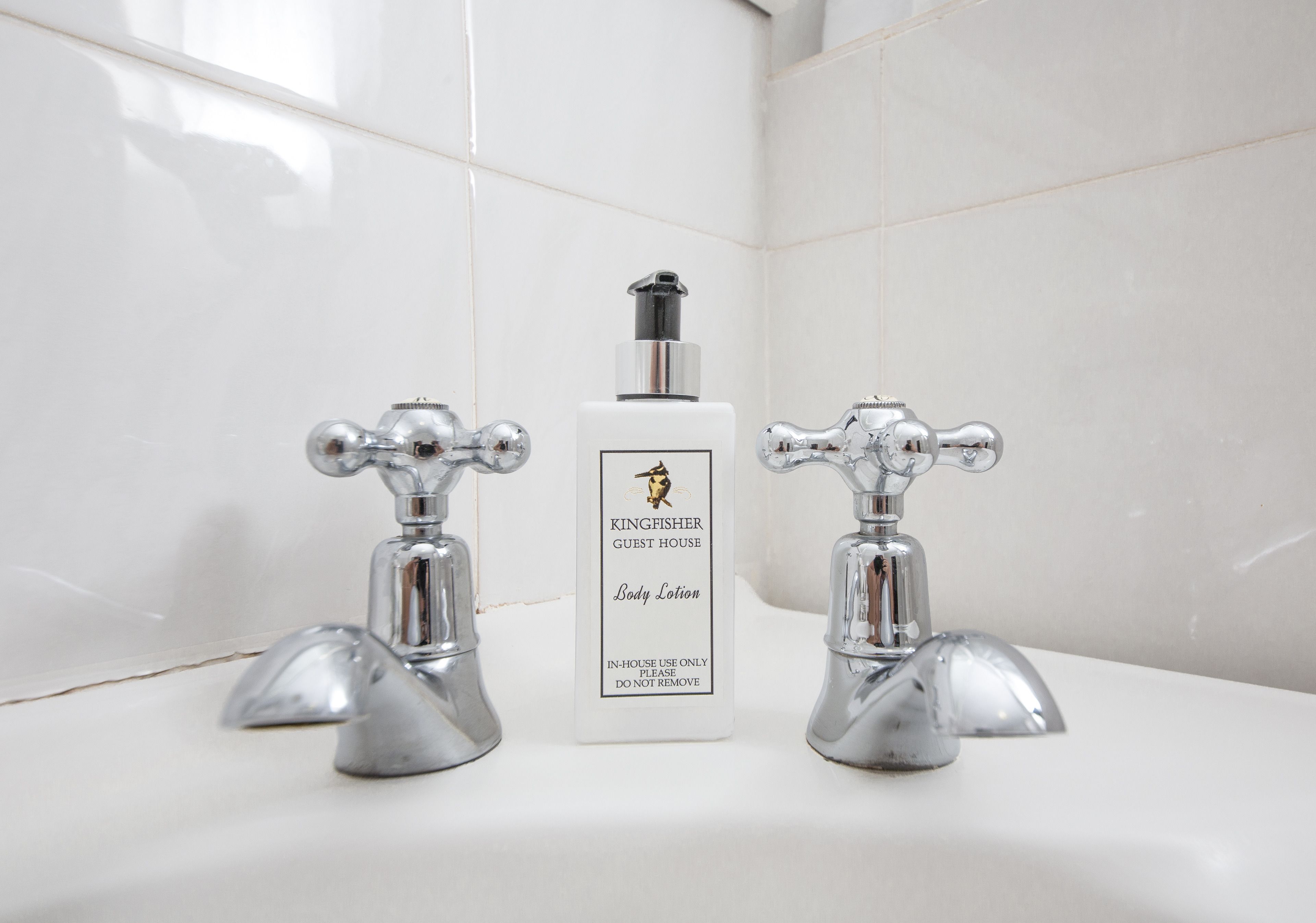 Ein Badezimmer mit zwei chromfarbenen Wasserhähnen und einer Flasche Körperlotion vom Kingfisher Guest House auf einem weißen Waschbecken.