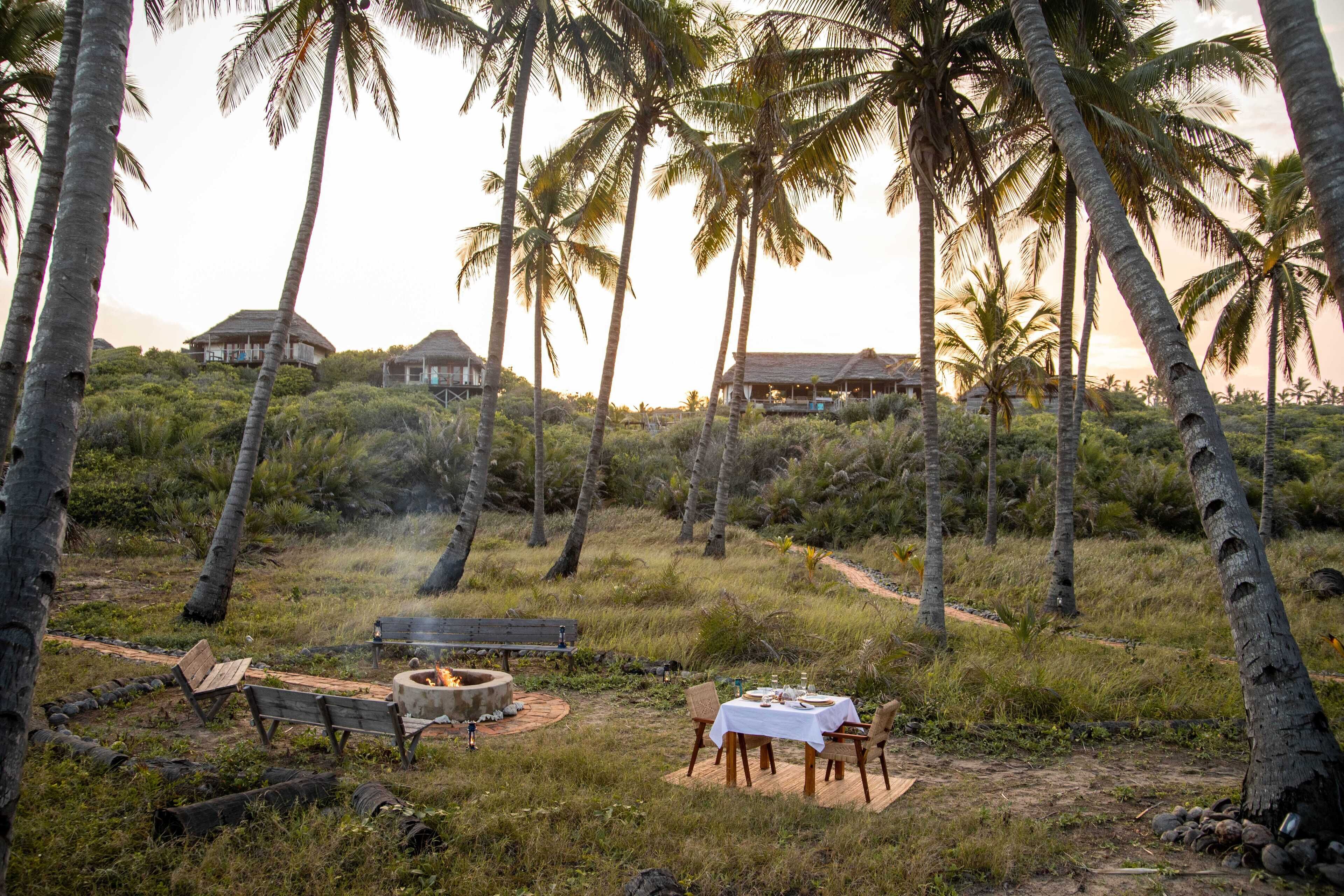 A tropical outdoor dining area with a fire pit, wooden benches, and a table set for two.