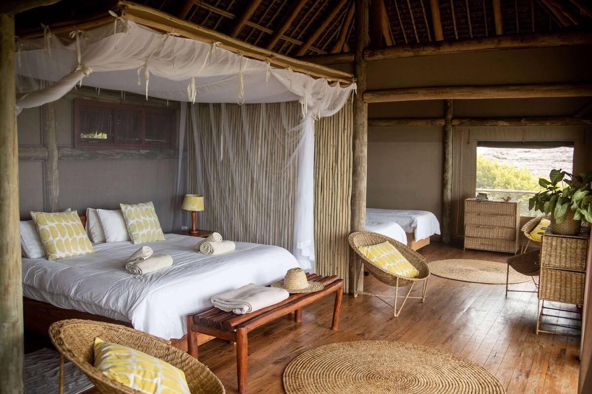 A bedroom with a canopy bed, a sitting area with wicker furniture, and a view of the outdoors.