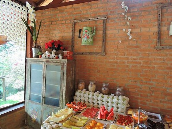 Un bufé de desayuno con diversas frutas y bollería, una pared de ladrillo rústica y un jarrón decorativo con flores.