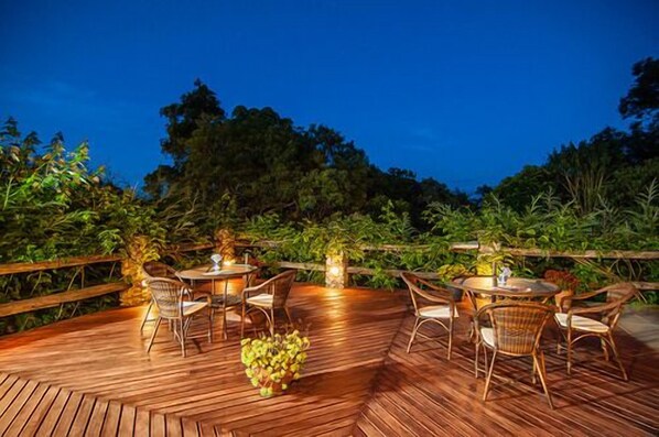 An outdoor dining area with wooden flooring and round tables, surrounded by chairs and greenery. - Pousada Cantinho das Estrelas (Camanducaia)
