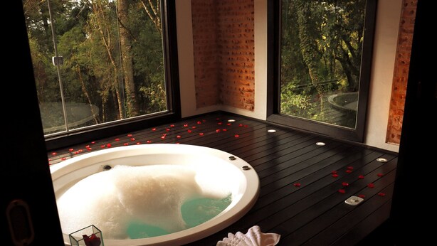 A spa room with a hot tub, wooden flooring, and large windows offering a view of trees. - Pousada Cantinho das Estrelas (Camanducaia)