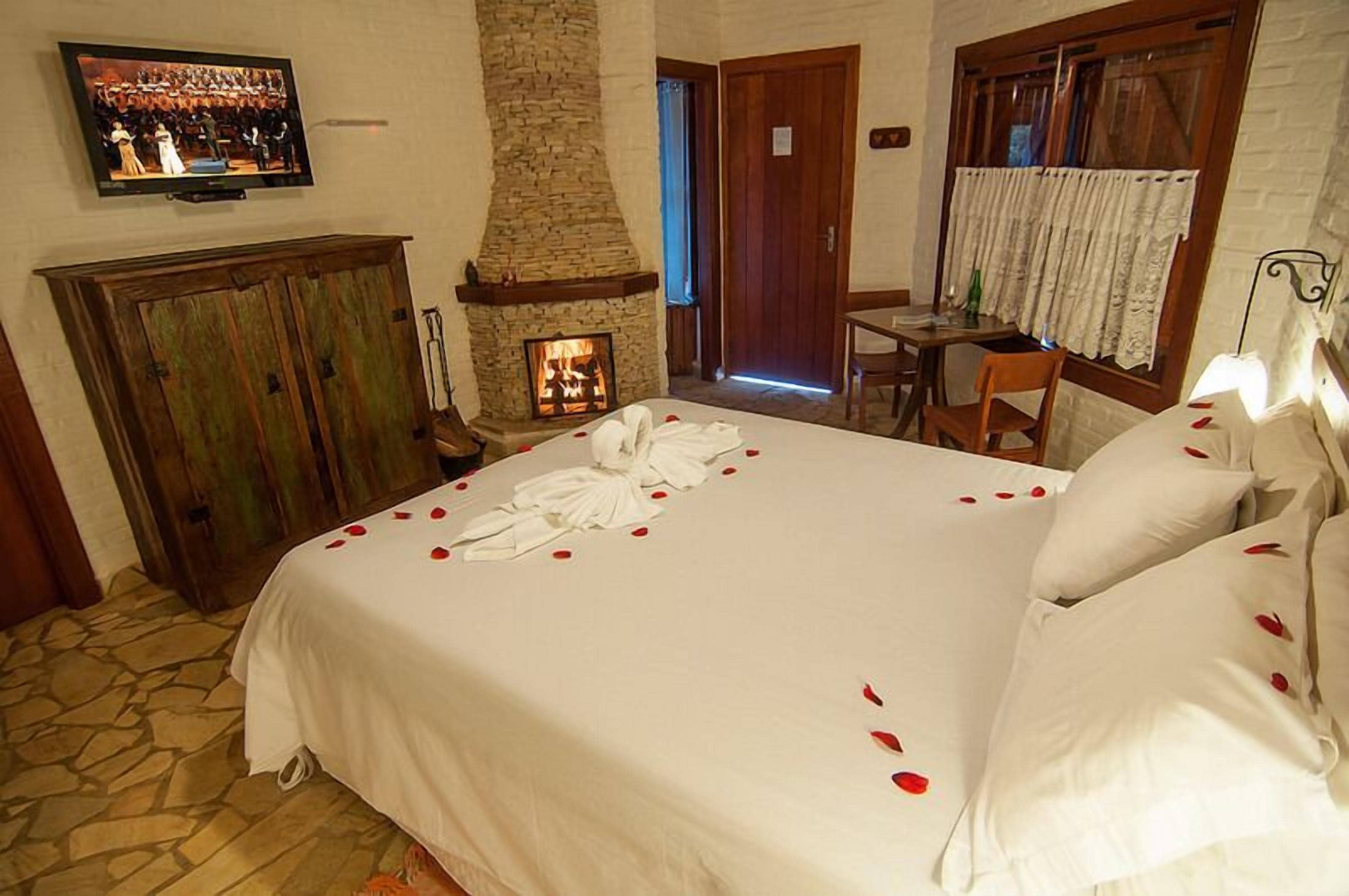 Une chambre d’hôtel avec une cheminée en pierre, un lit parsemé de pétales de rose, un téléviseur fixé au mur et une armoire en bois.