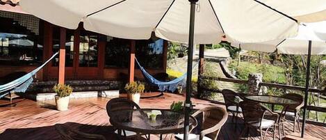 Un patio al aire libre con piso de madera, mesas, sillas y una sombrilla grande que proporciona sombra.