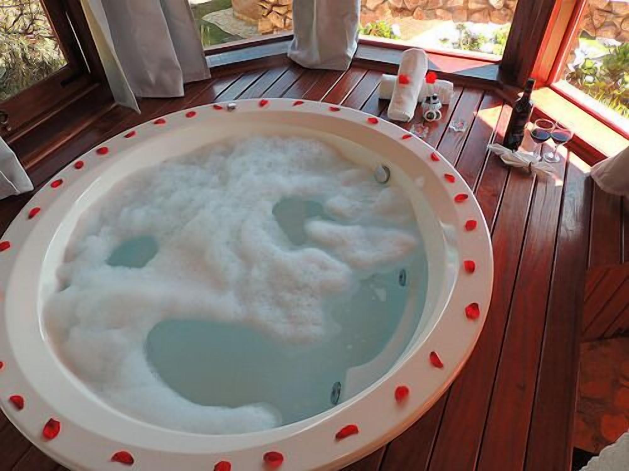 A hot tub with rose petals on the edge, surrounded by wooden decking and a window with curtains.