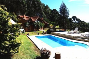 A swimming pool with a stone deck, surrounded by a grassy lawn and trees. - Pousada Cantinho das Estrelas (Camanducaia)